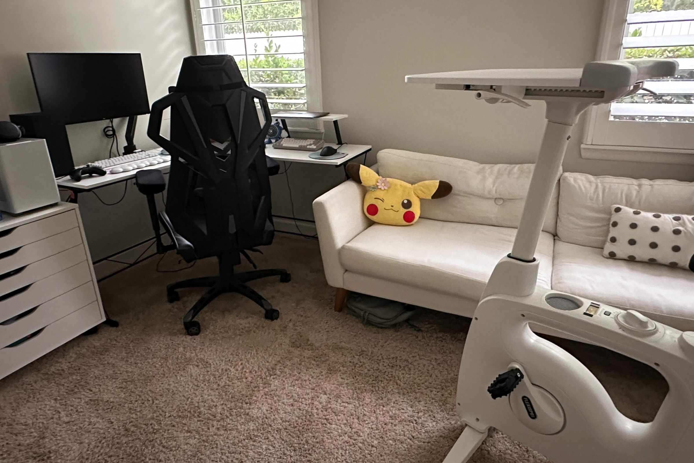 Part of home office space with desk, window, couch with Pikachu and cycling desk (an exercycle with a surface for a laptop)