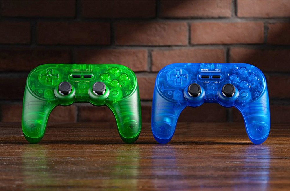 The new transparent blue and green 8BitDo Pro 2 controllers on a wooden desk against a brick wall.