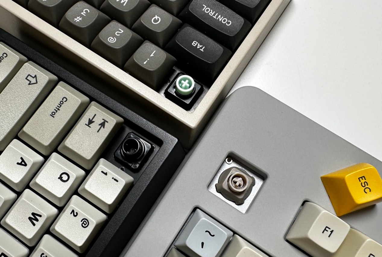 Close up shot of three keyboards with one keycap removed from each, showing the switch stems. Top (green) stem is MX-compatible Deskeys slider, center (beige) is the Norbauer Aerostem in a Seneca case, and on the lower left a black stock Topre stem.