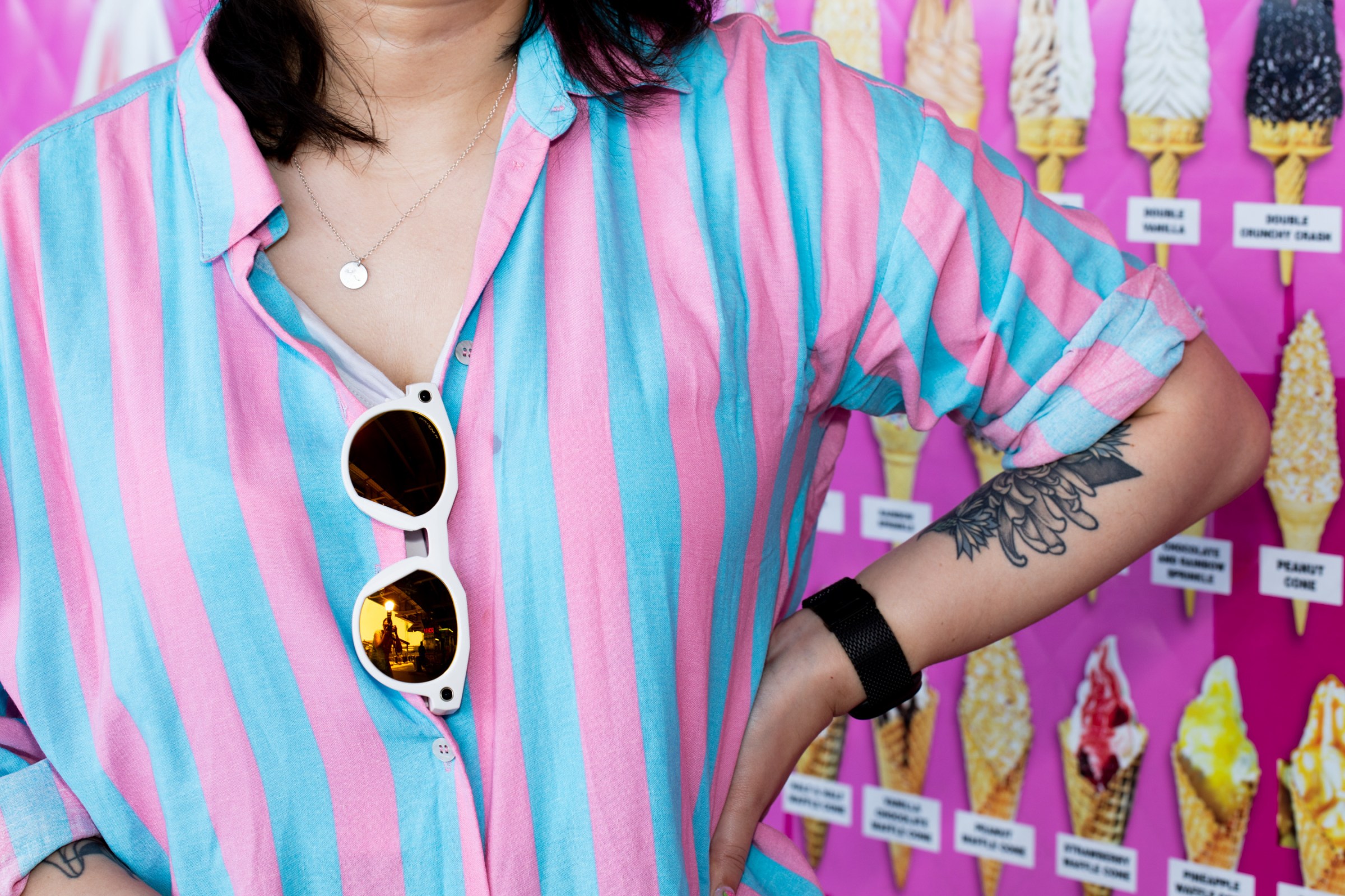Wide angle view of the Oakley Meta glasses slung in Senior Reviewer Victoria Song’s shirt, which is striped blue and pink. There is a colorful pink ice cream truck menu in the background.