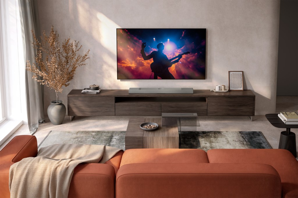 A living room setup showing a KEF XIO soundbar positioned on a unit below a wall-mounted TV. There is an orange sofa in the foreground and a coffee table in between.