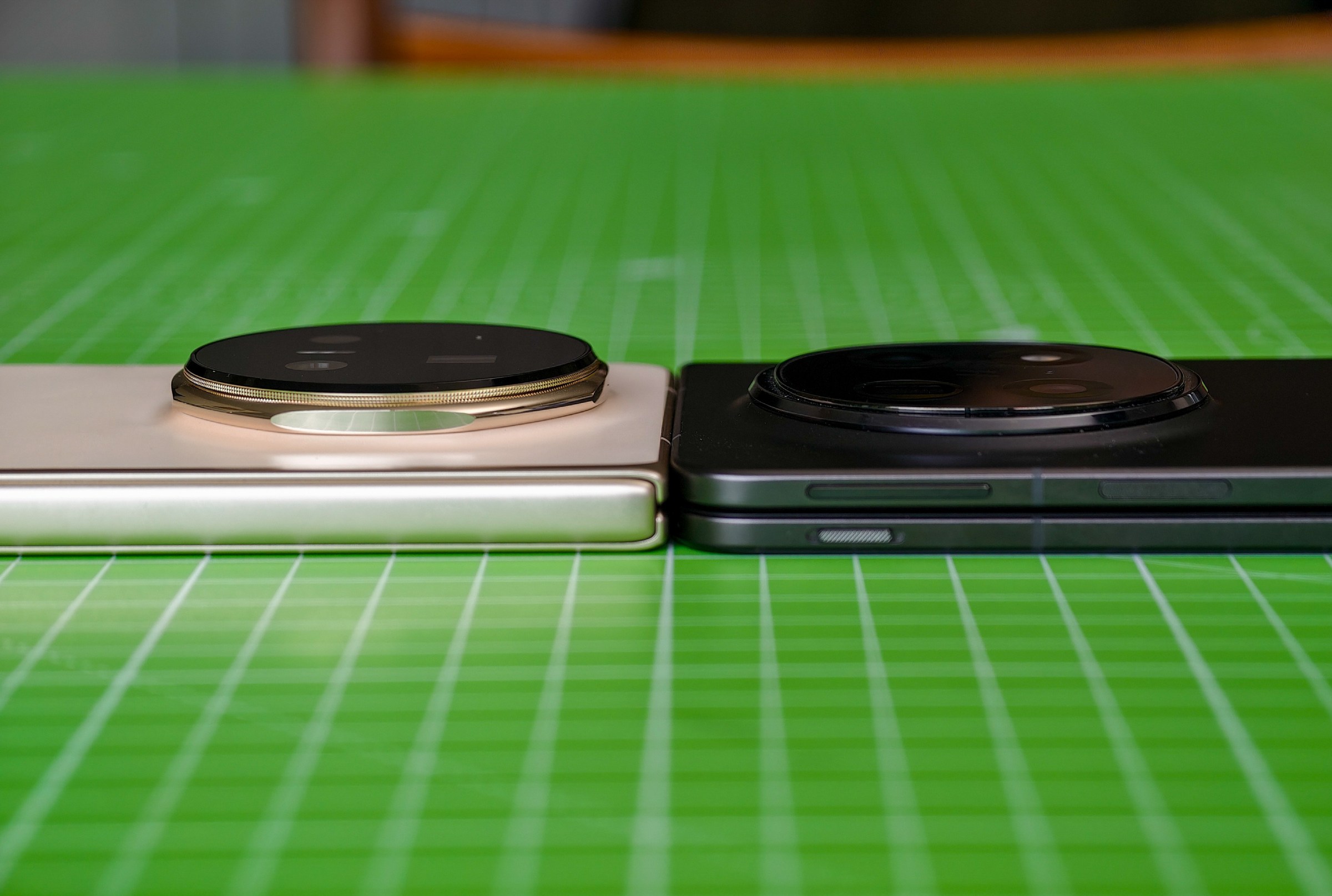 The Honor Magic V5 and Oppo Find N5 both lying closed on a green cutting mat, showing that the Honor’s camera bump is visibly taller