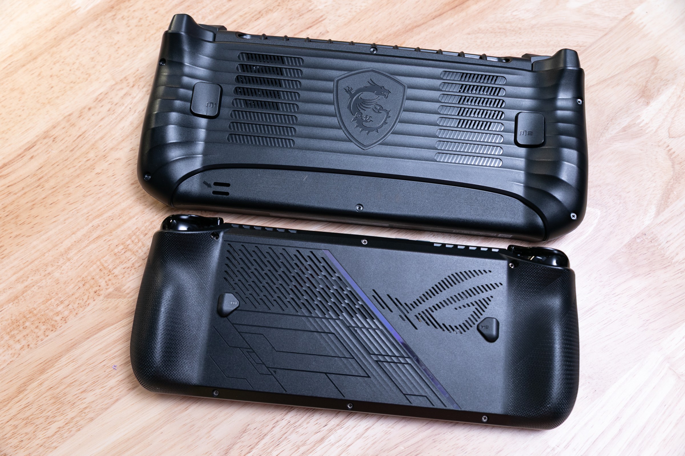 The backs of the MSI Claw 8 AI Plus and Asus ROG Ally X, two black handhelds, to show size and grip comparison. The Ally X is shorter and more squat.