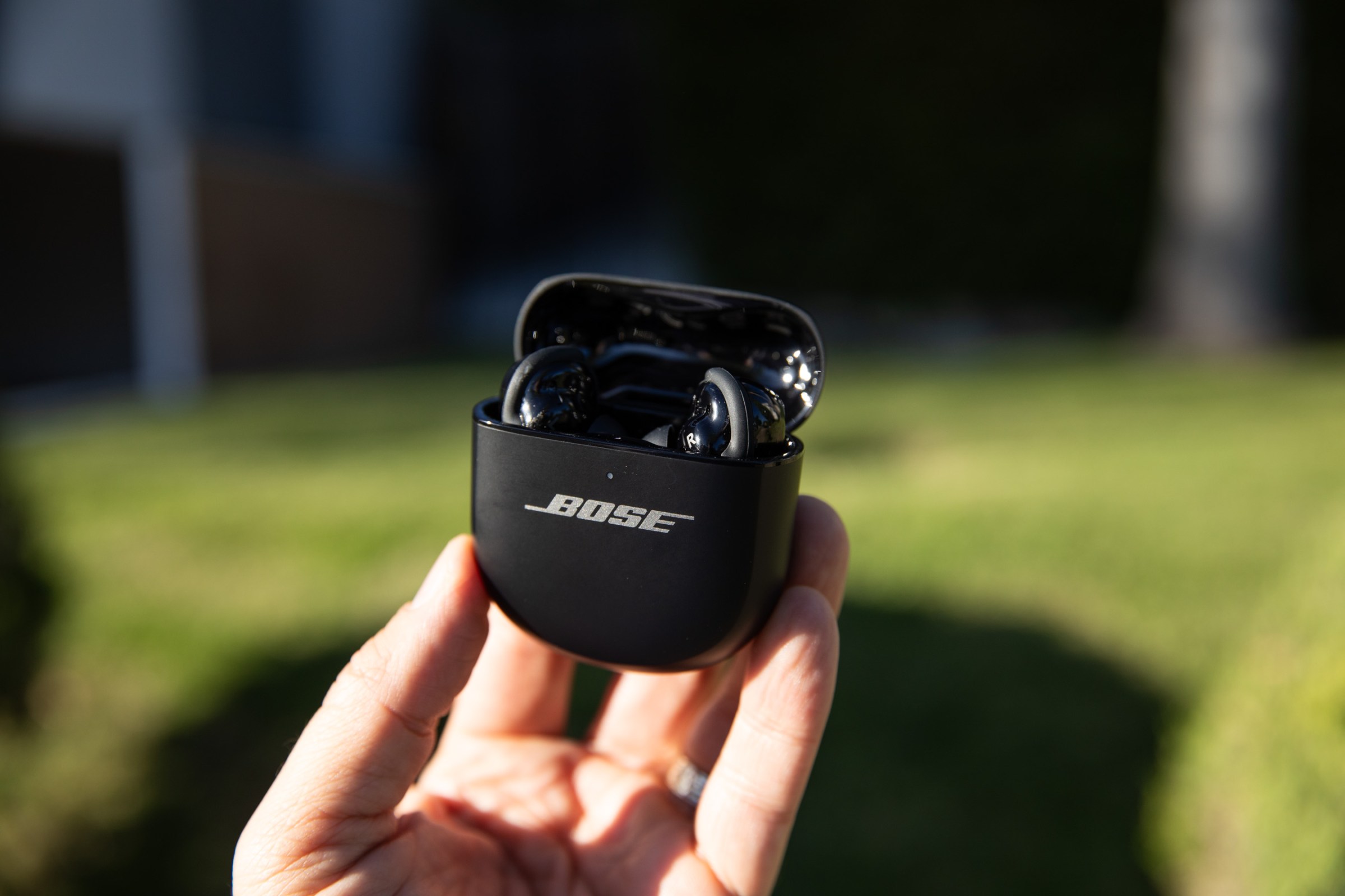 A hand holding the Bose Ultra Earbuds Gen 2 in their open charging case, outside in front of some grass.