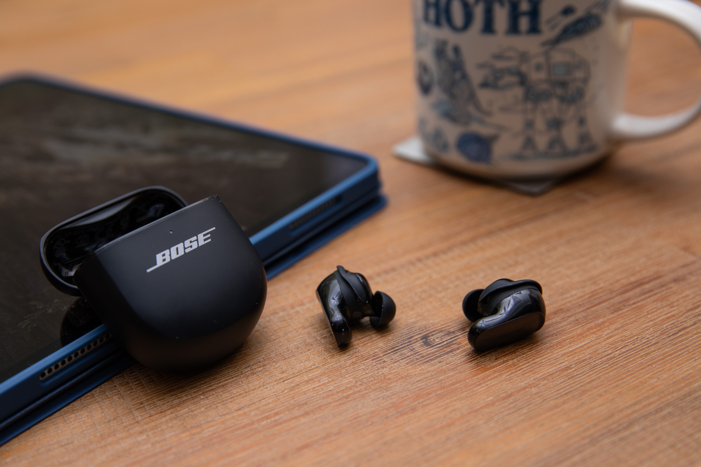 The Bose Ultra gen 2 earbuds on a kitchen table with their case leaning against an iPad and a Star Wars coffee mug in the background.