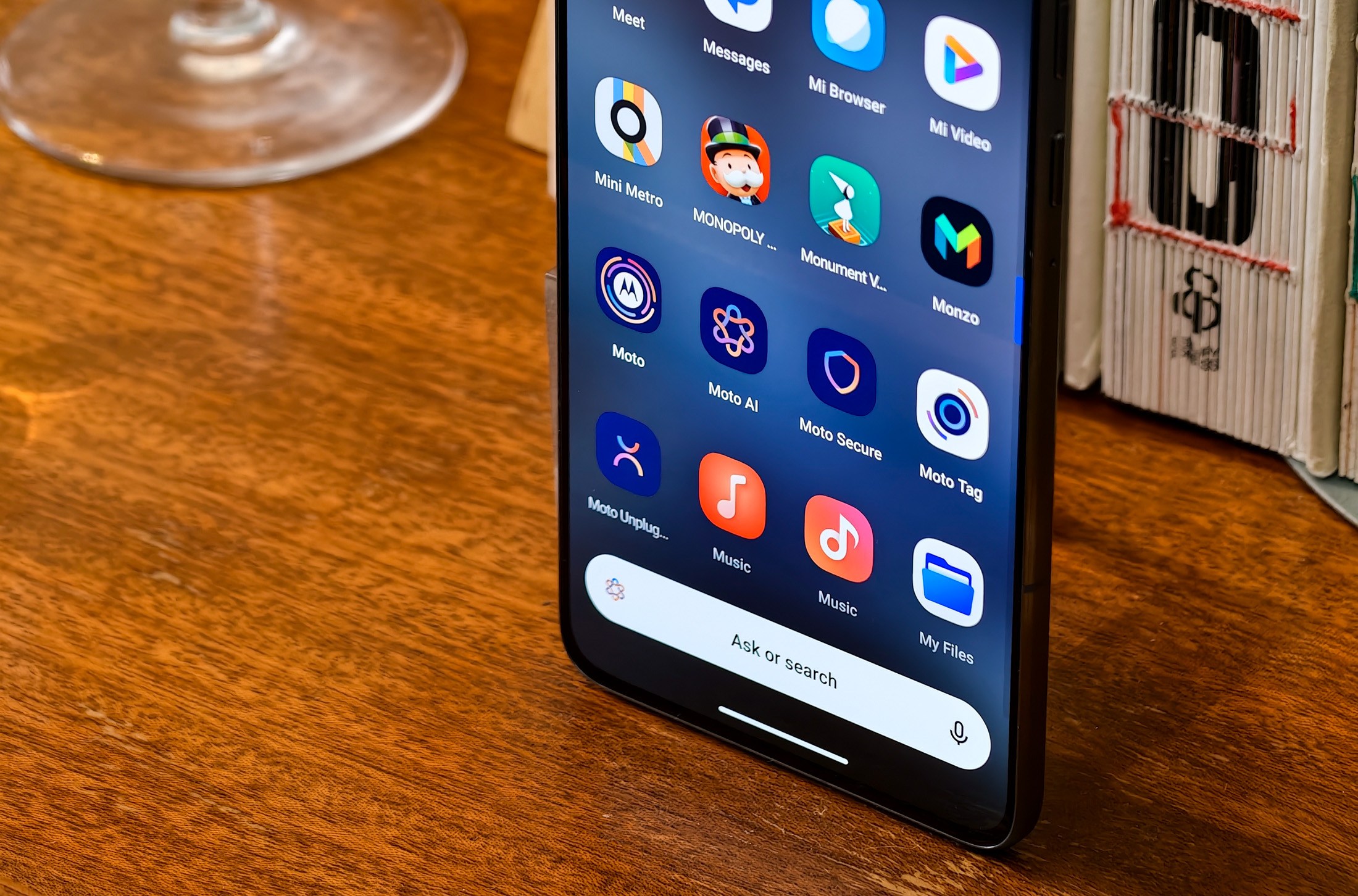 Photo of Motorola Edge 70 standing on a wooden table in front of some glasses, showing the app drawer with preinstalled Motorola apps