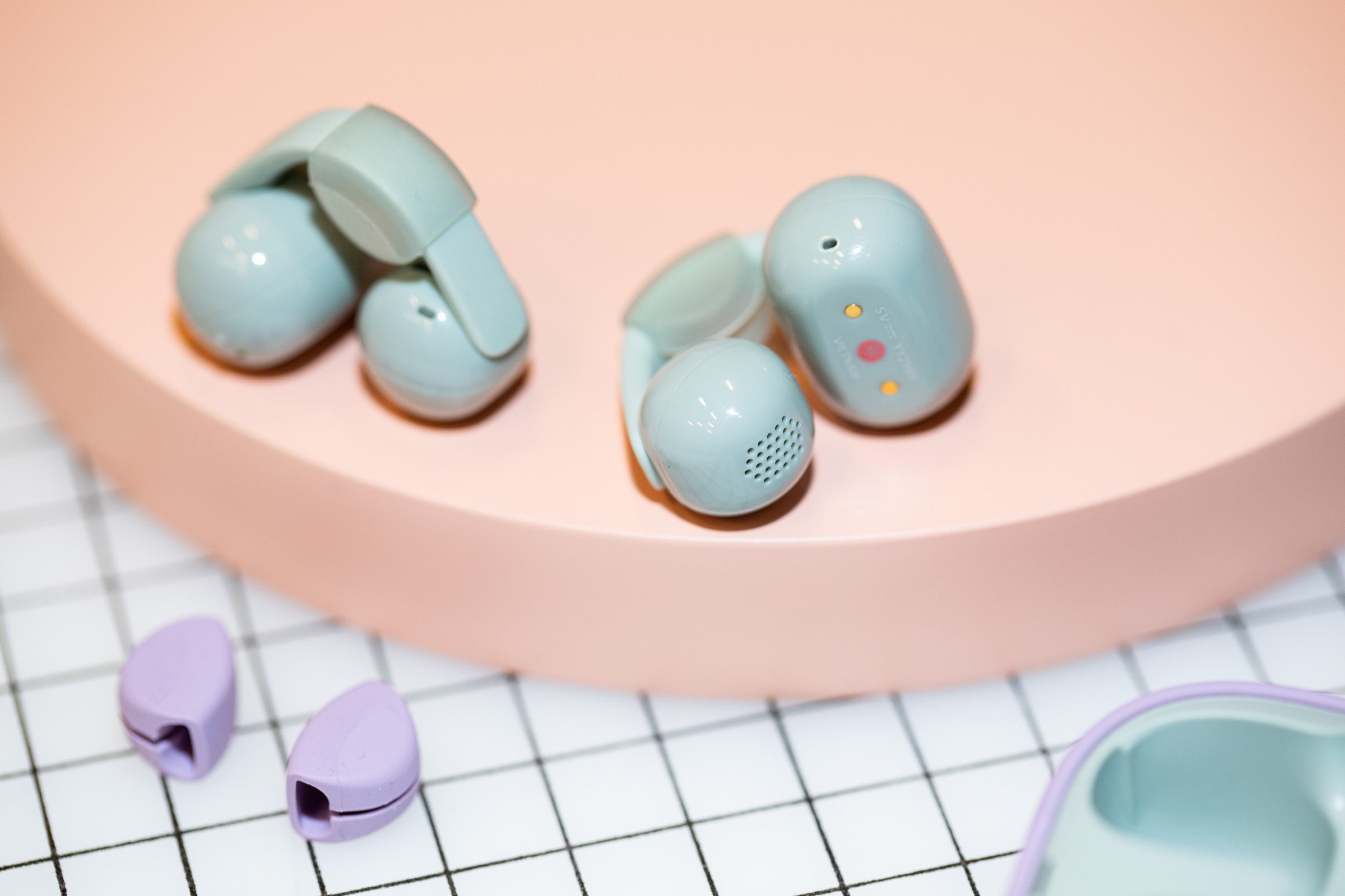 The Sony LinkBuds Clip earbuds with their clear silicone cushions on a peach circular surface above lavender cushions on a graph paper surface.