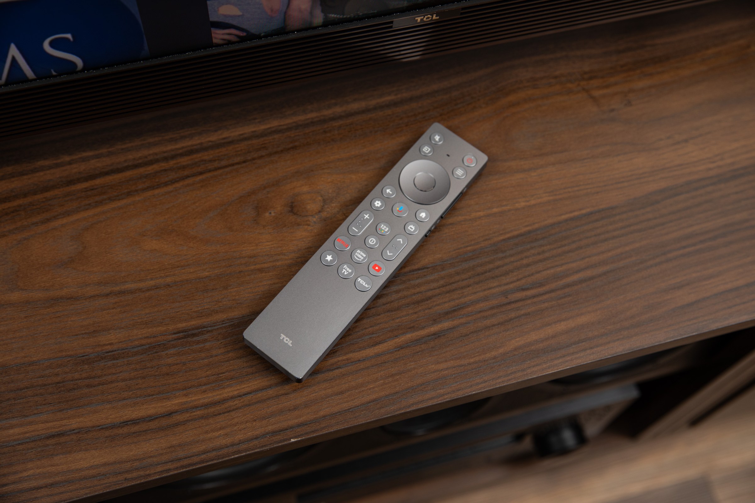 The TCL X11L remote on a dark wood TV table.