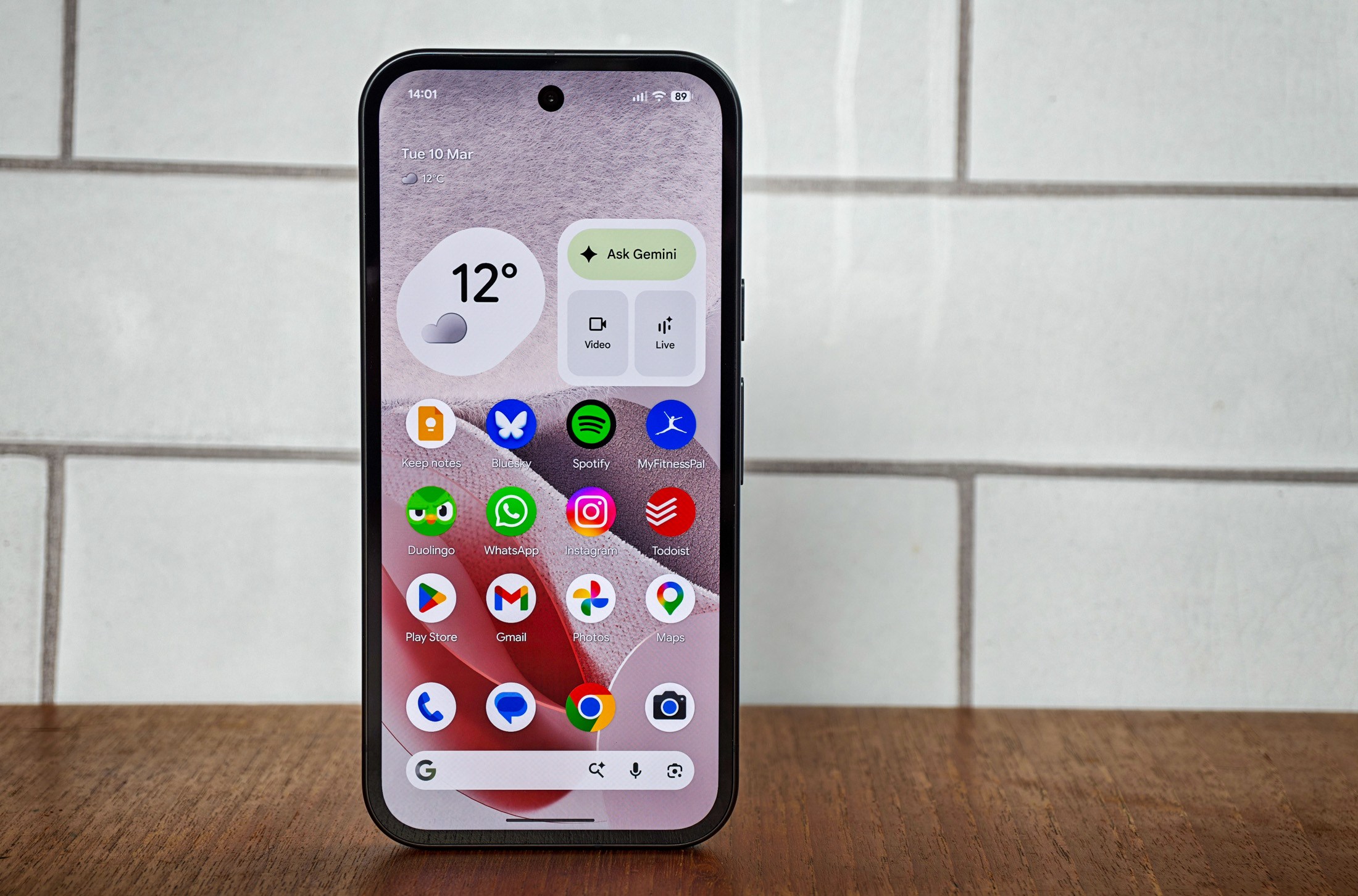 Photo of Google Pixel 10A in front of white tiles, showing the home screen