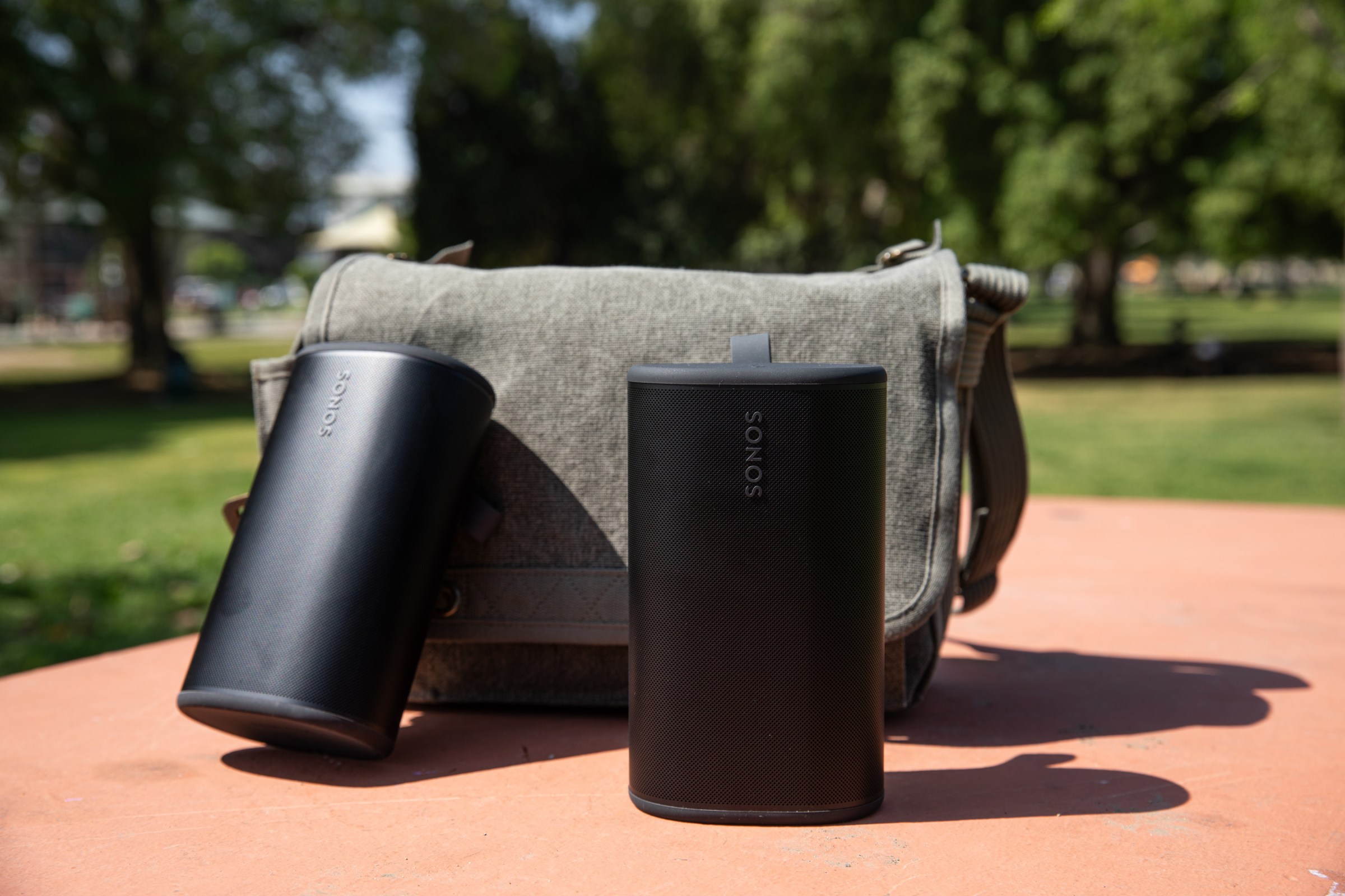 Two Sonos Play speakers in front of a sage green bag on a pink colored park table.