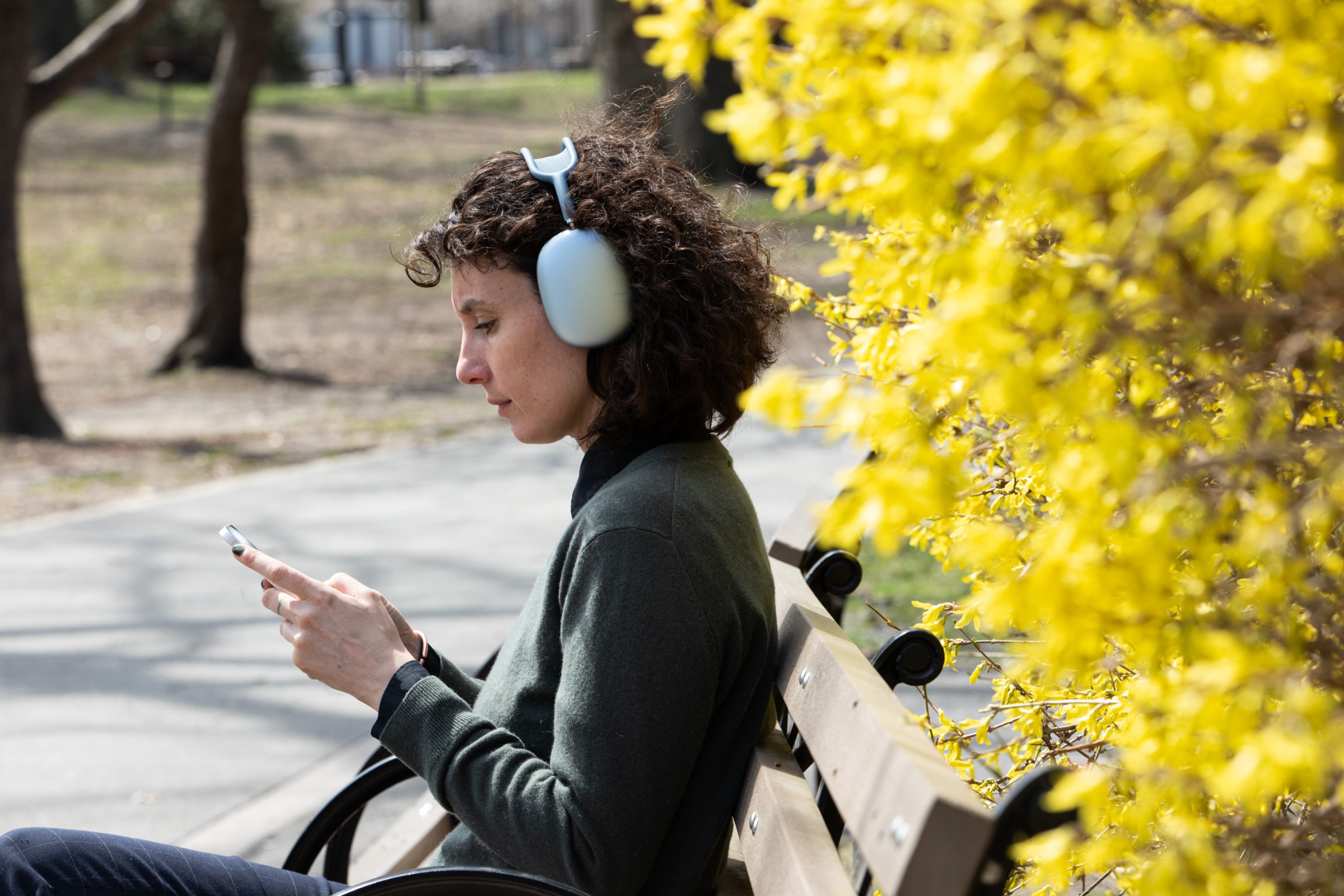 A person sitting on a bench wearing the blue Apple AirPods Max 2 while looking at their phone.