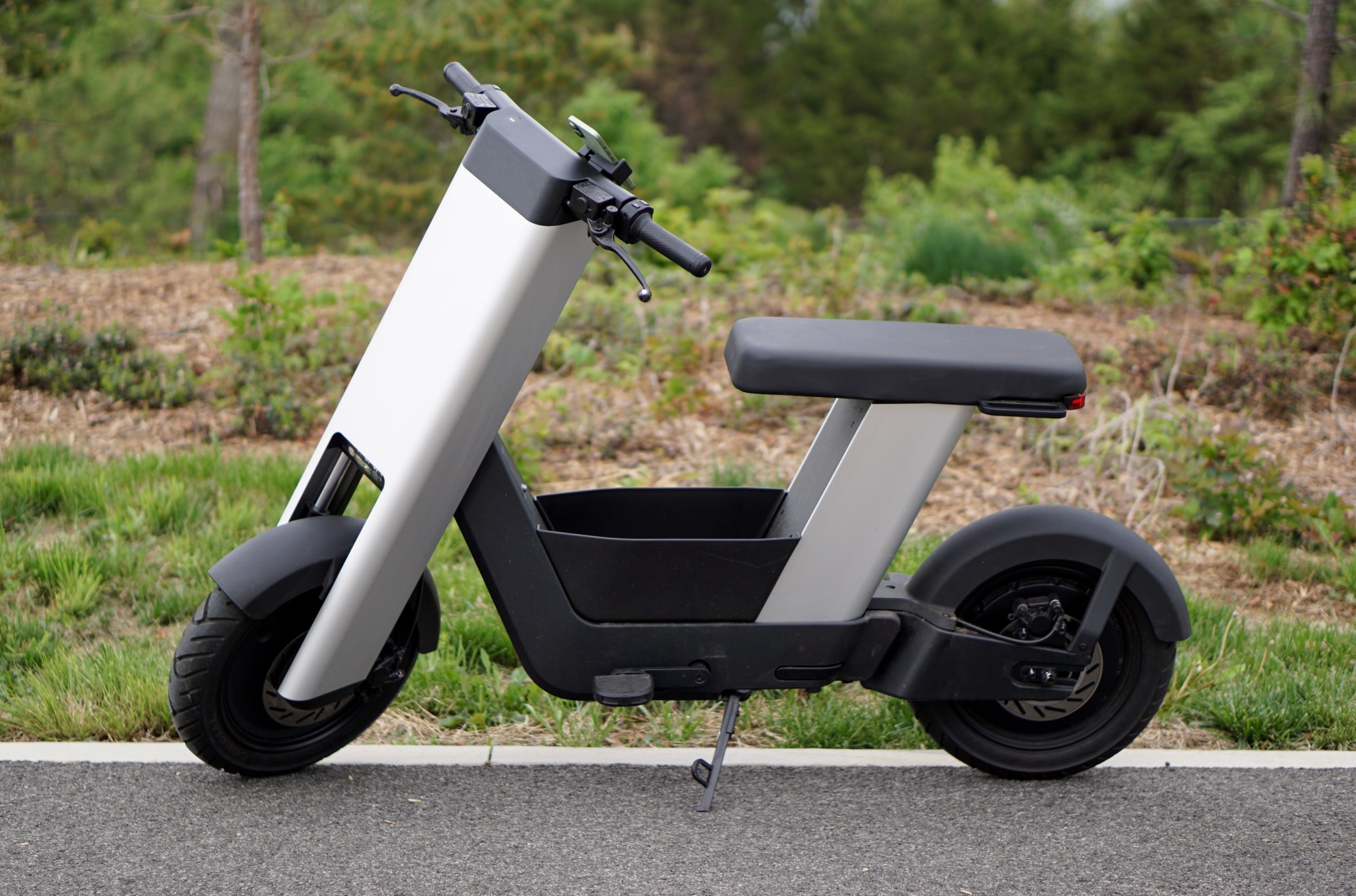 A photo of a silver e-bike from the side, in front of trees.