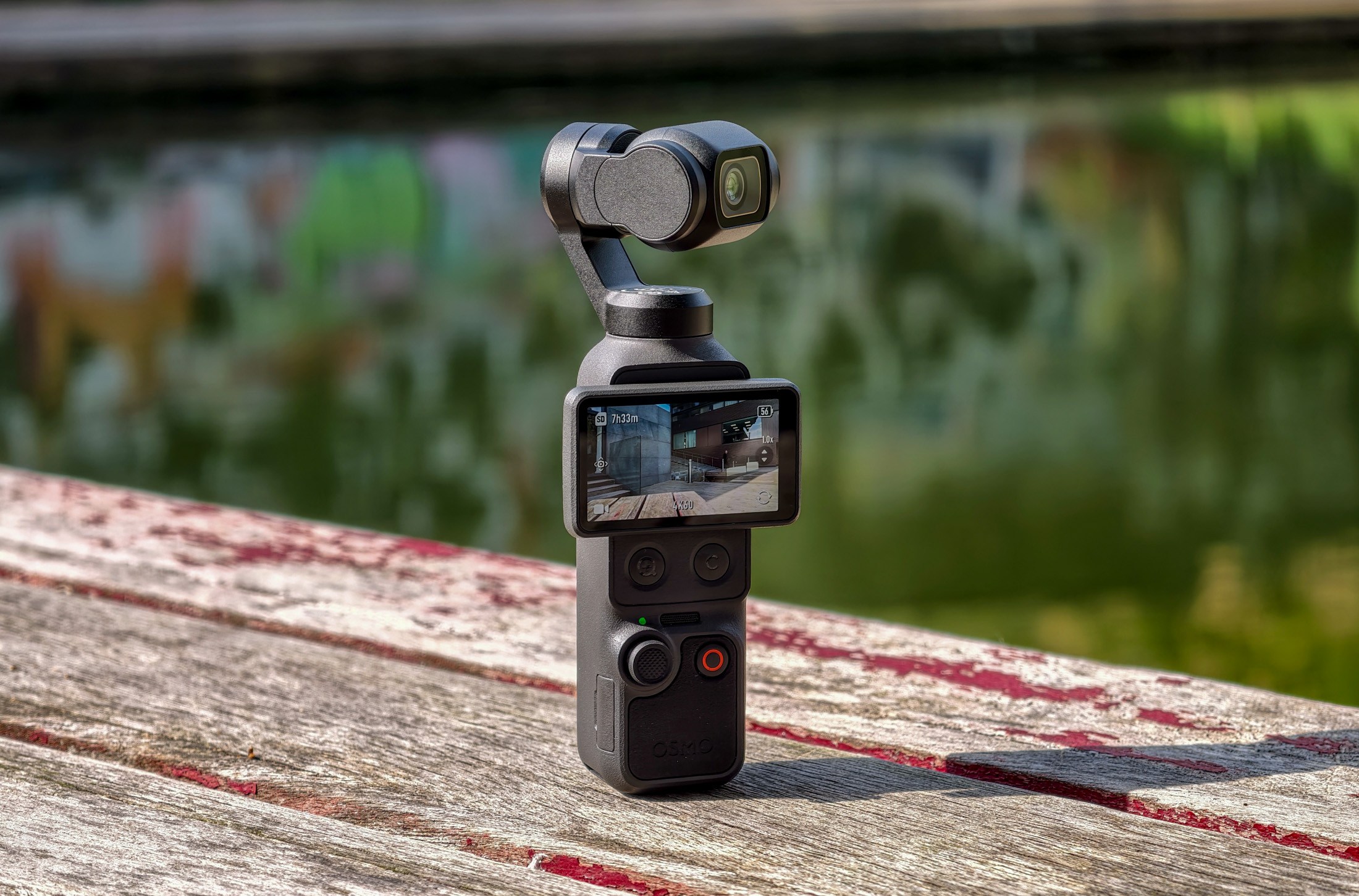 Photo of the DJI Osmo Pocket 4 standing on a wooden table in front of a canal, showing the screen with the camera facing off at an angle