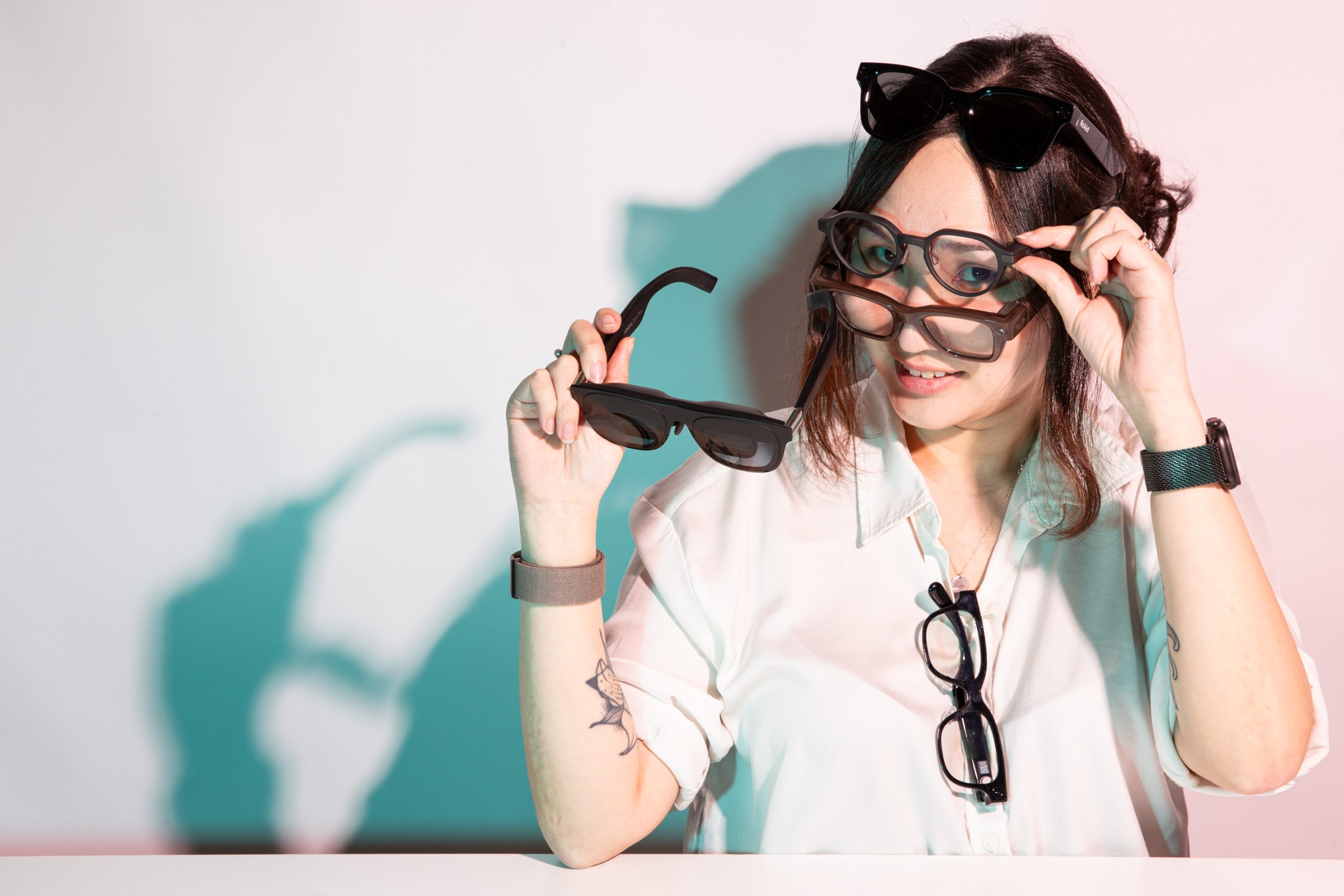 Senior reviewer Victoria Song wearing three pairs of smart glasses while holding a fourth and hanging a fifth from her collar. The expression is cheeky and reminiscent of Risky Business movie poster.