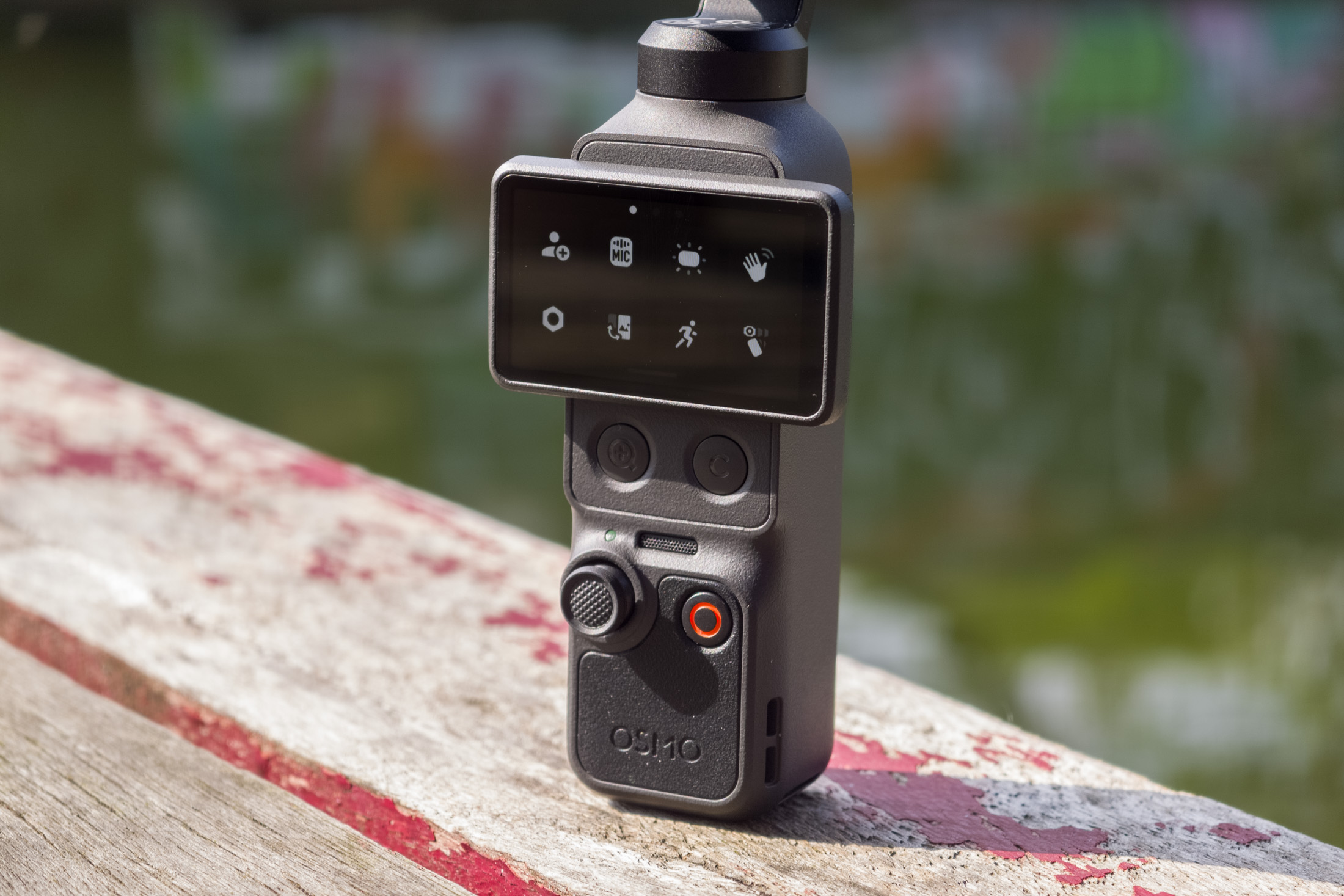 Photo of the DJI Osmo Pocket 4 standing on a wooden table in front of a canal, showing the screen with the settings menu