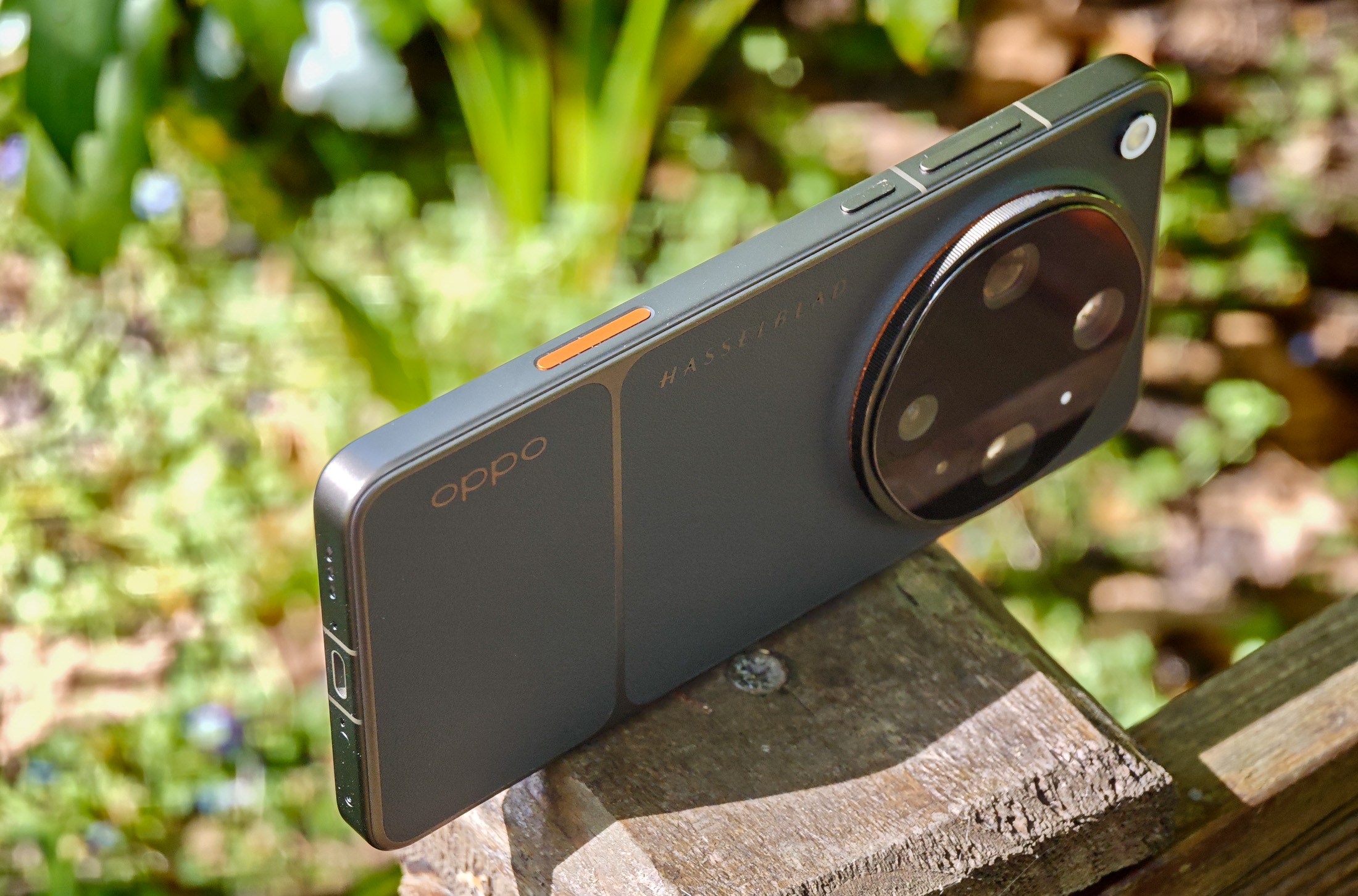 Photo of Oppo Find X9 Ultra resting horizontally on a wooden post in front of green plants, showing the orange shutter button