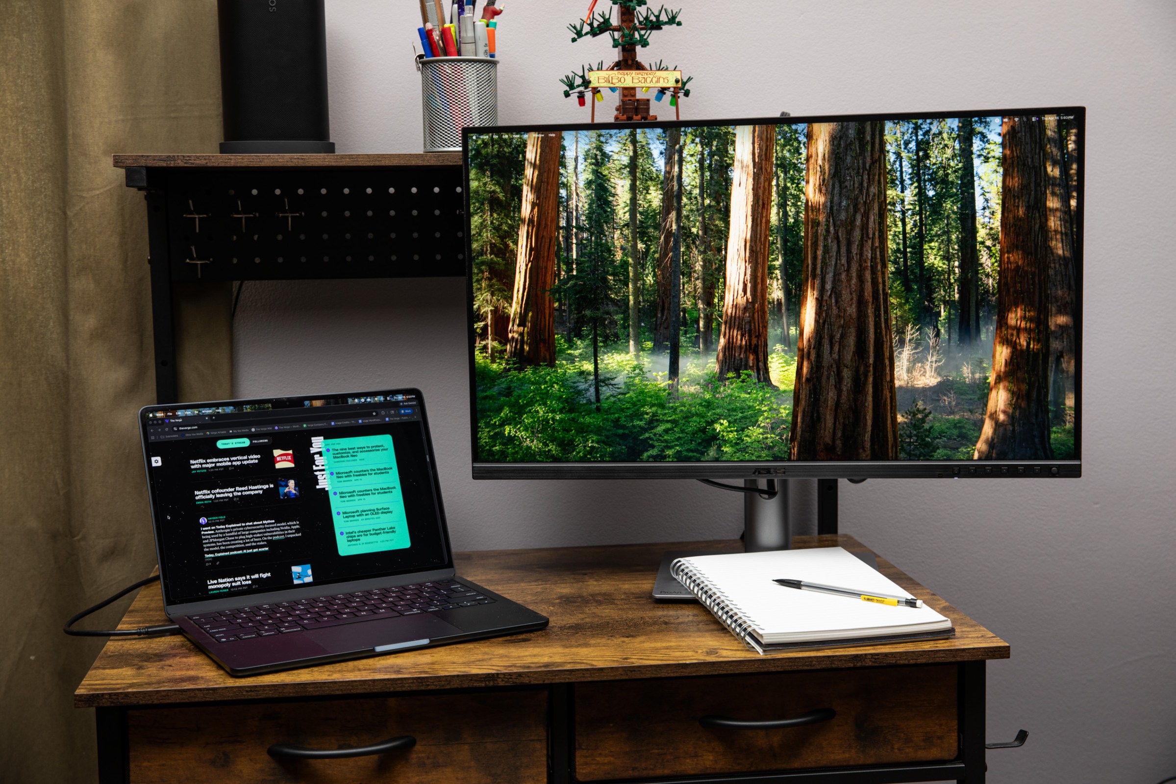 The Asus ProArt PA27JCV 5K monitor on a wooden desk next to a MacBook with a notepad and pen in front of it.