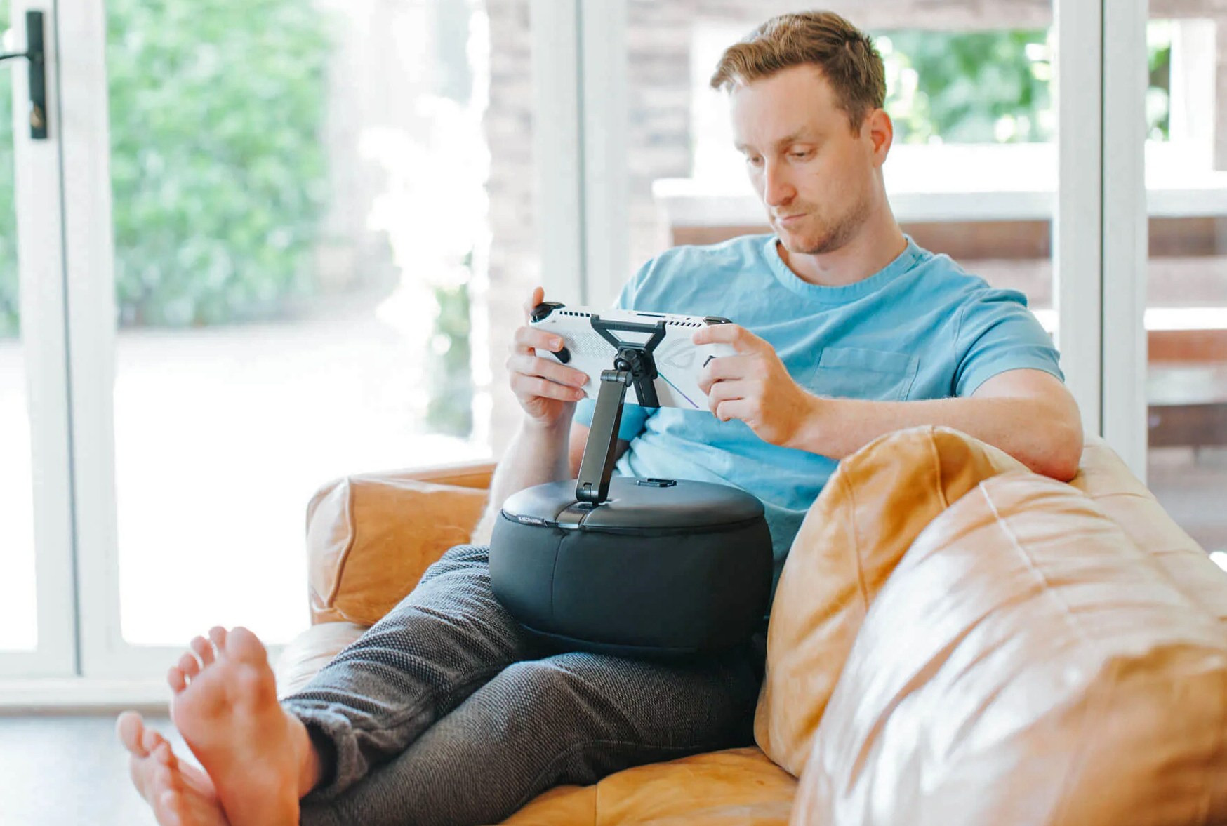 A gamer plays a handheld console on a sofa attached to the Gaming Pillow on their lap.