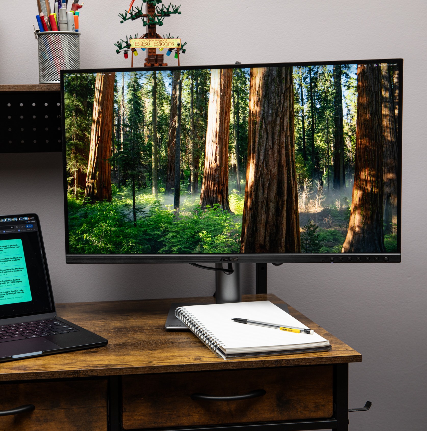 The Asus ProArt PA27JCV 5K monitor on a wooden desk next to a MacBook with a notepad and pen in front of it.