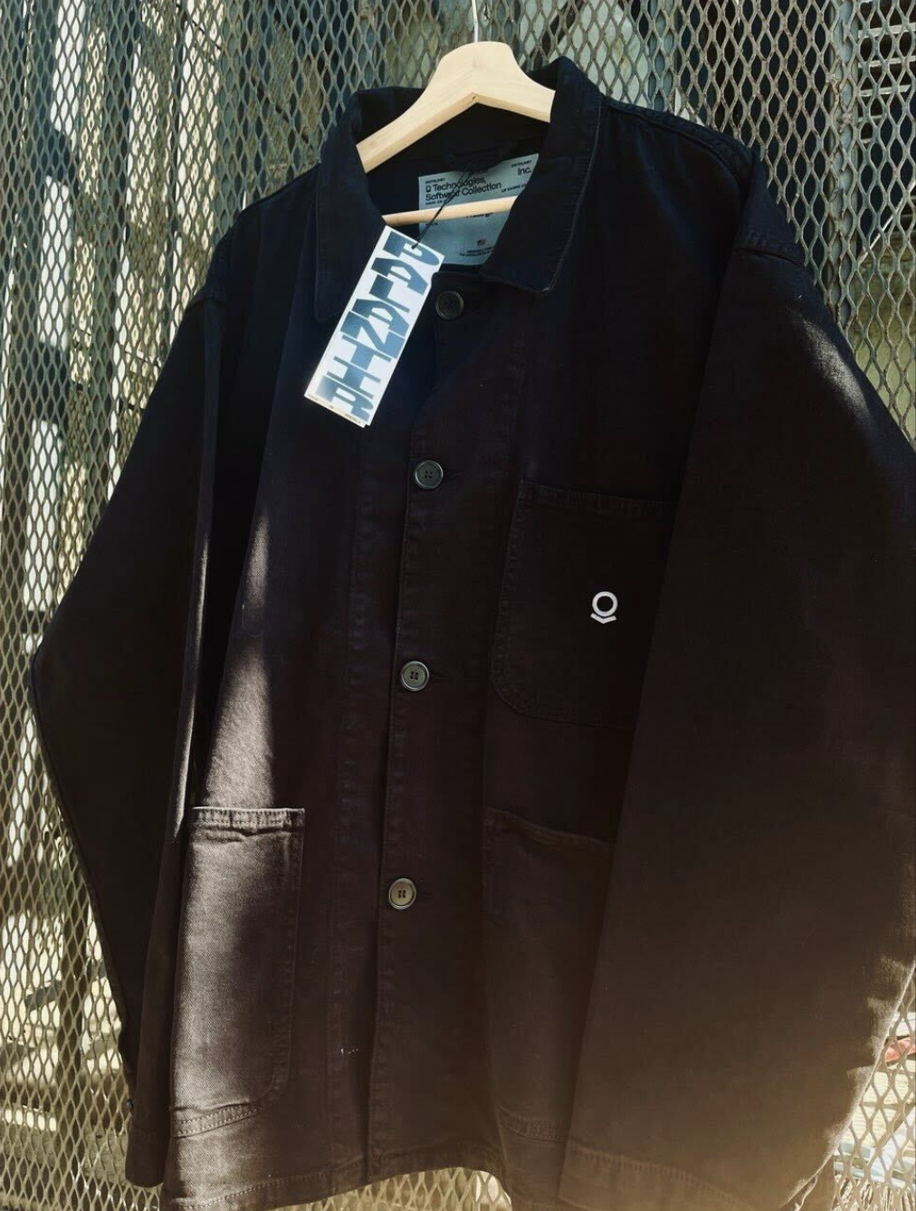 A black Palantir-branded chore coat hanging on a hanger.