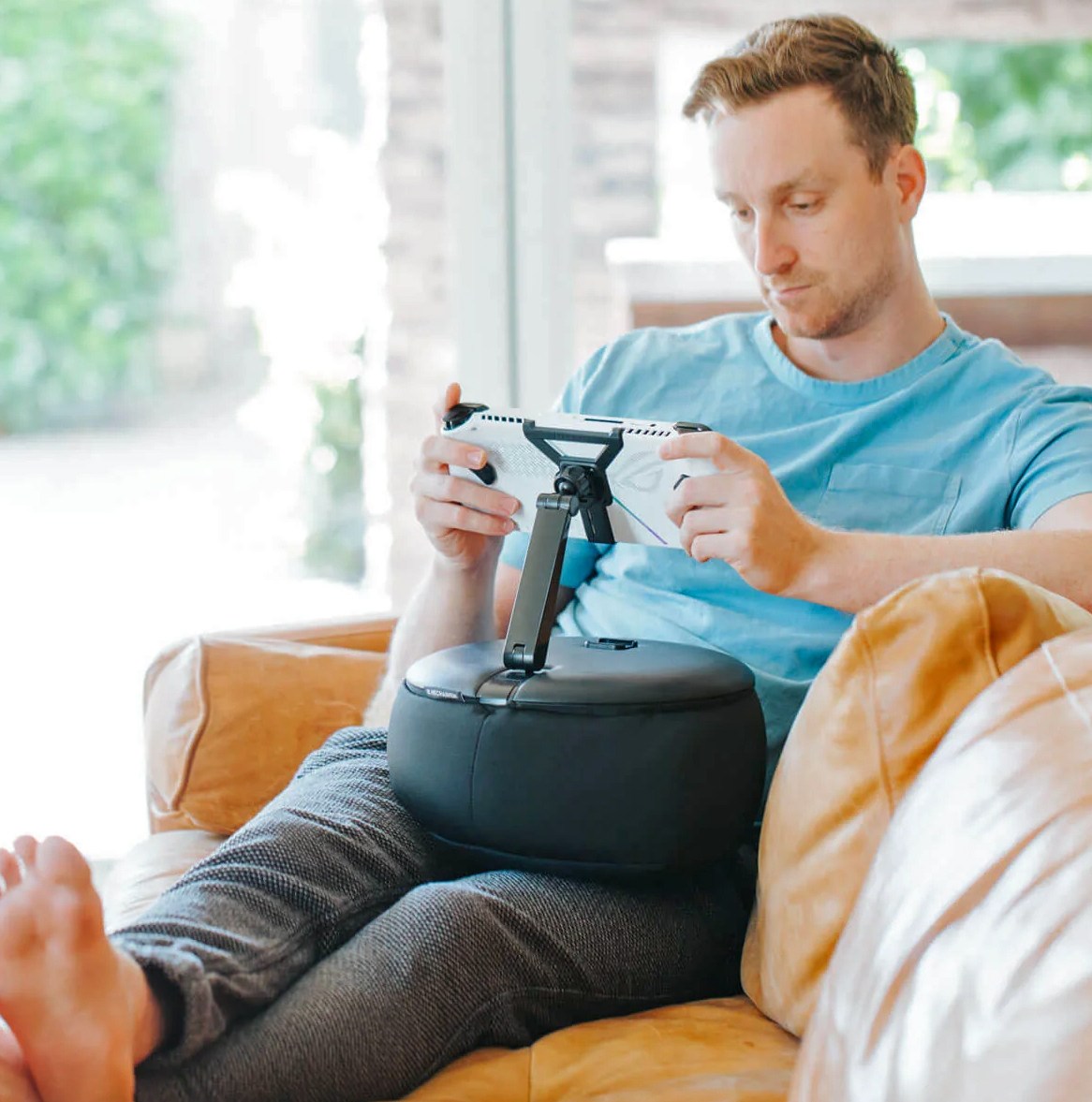 A gamer plays a handheld console on a sofa attached to the Gaming Pillow on their lap.