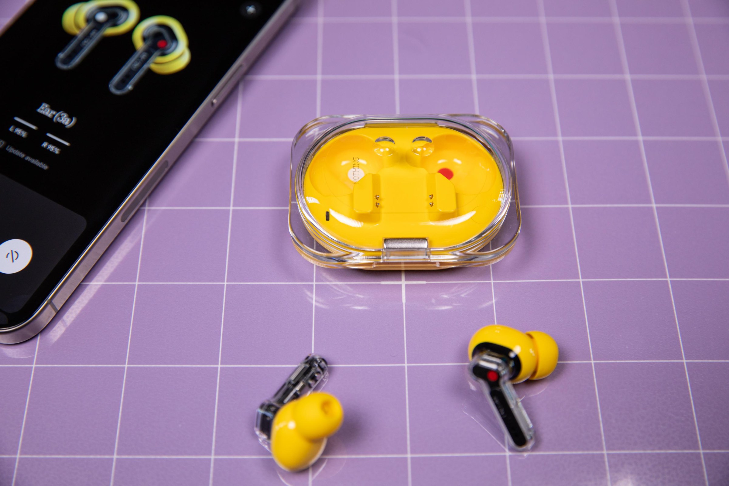 The yellow Nothing Ear 3A earbuds and case on a light purple grid background with a iPhone nearby.