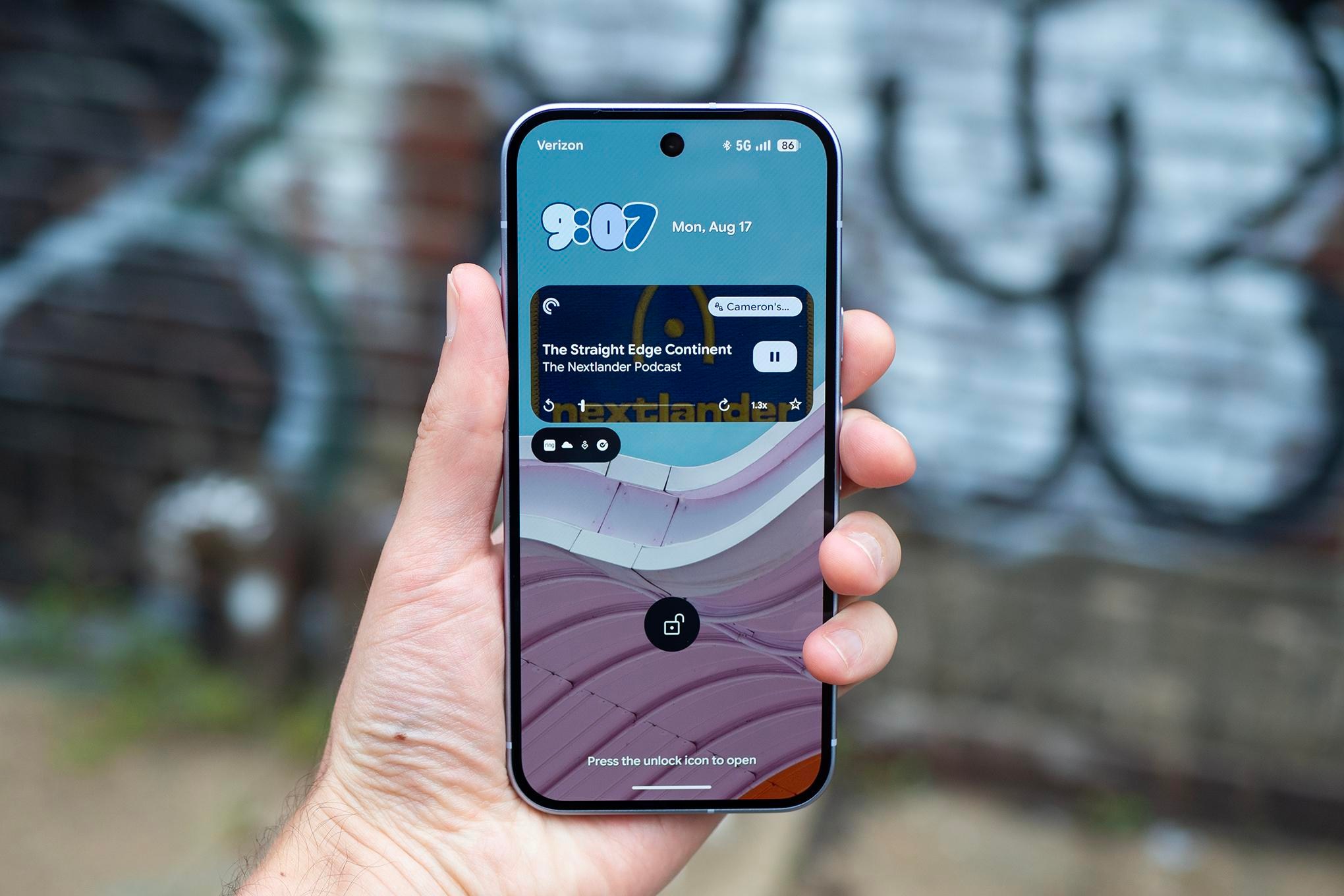 Illuminated screen of a Google Pixel 11 in a person’s hand in front of a blurry background of graffiti.