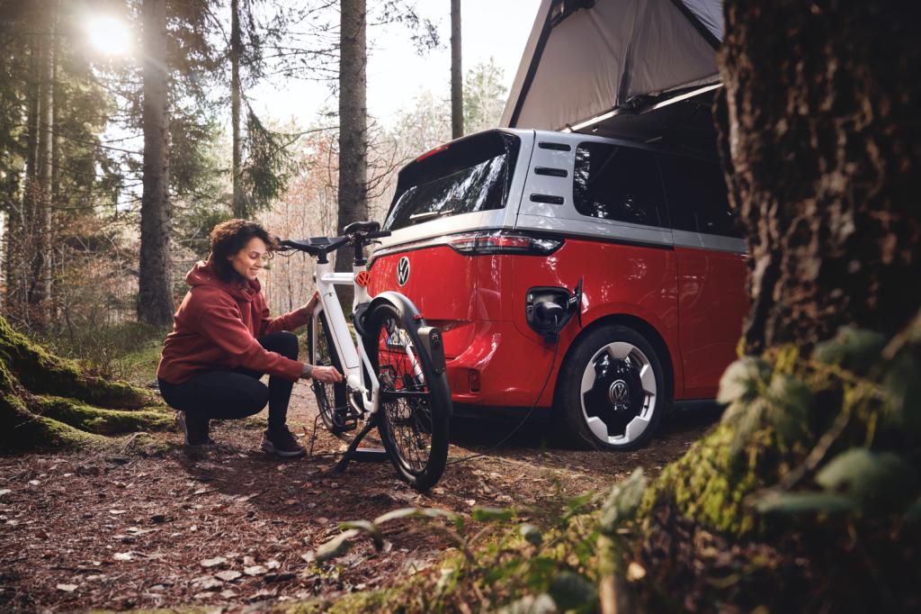 A Volkswagen Smart eBike in a forest, plugged into a parked VW car. A woman is kneeling next to it and attending to the pedals.