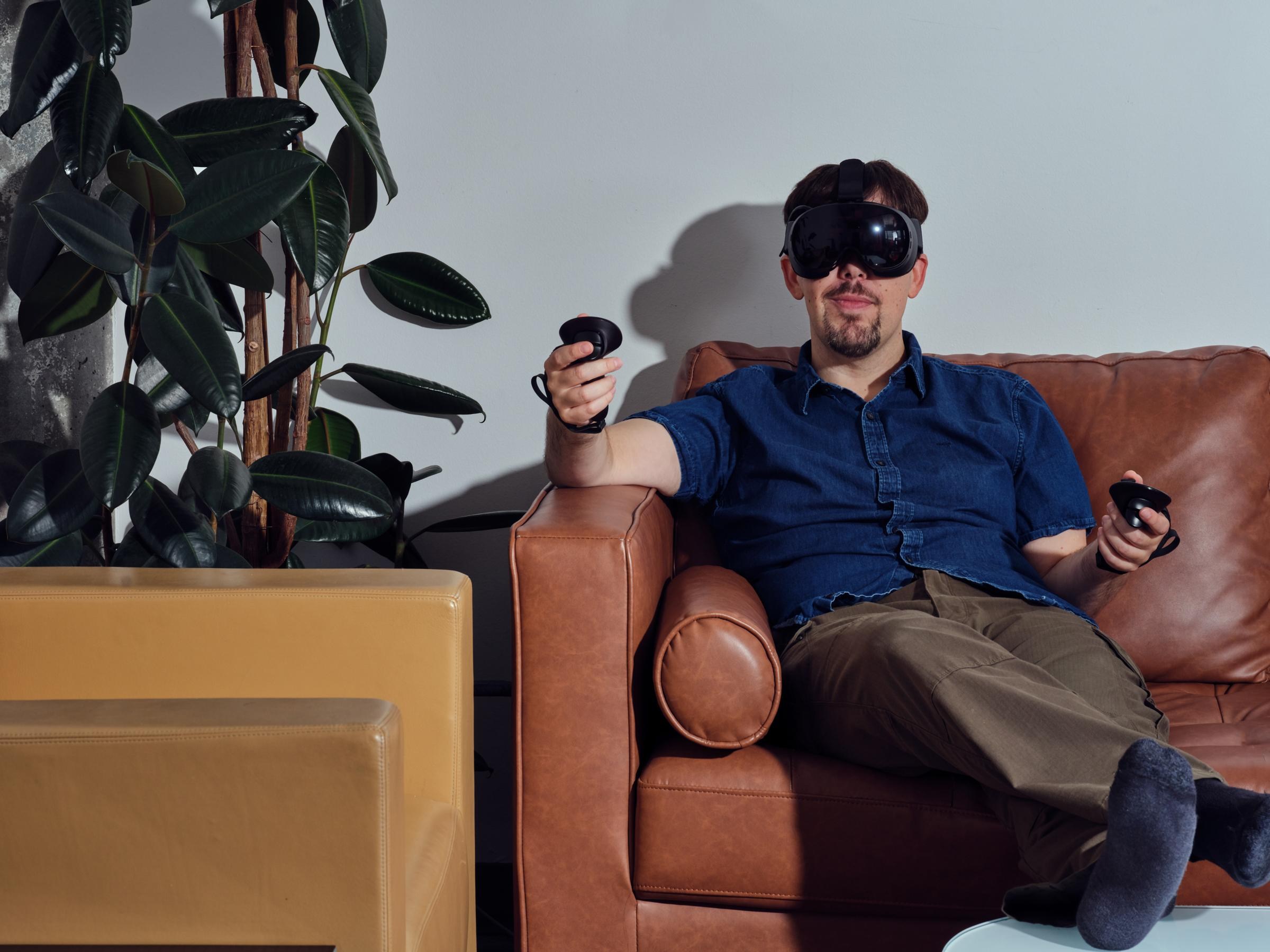 Sean lounges on a couch facing the camera while inside the Steam Frame, with one wand-holding arm propped up on an arm rest and the other against the rear cushion. A rubbery leaf plant is visible to his right.
