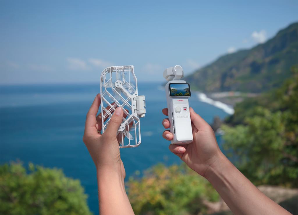 Somebody holding the two parts of a HoverAir Versa. In the left hand is the Flight Kit, and in the right is the camera itself. The sea is visible in the background.