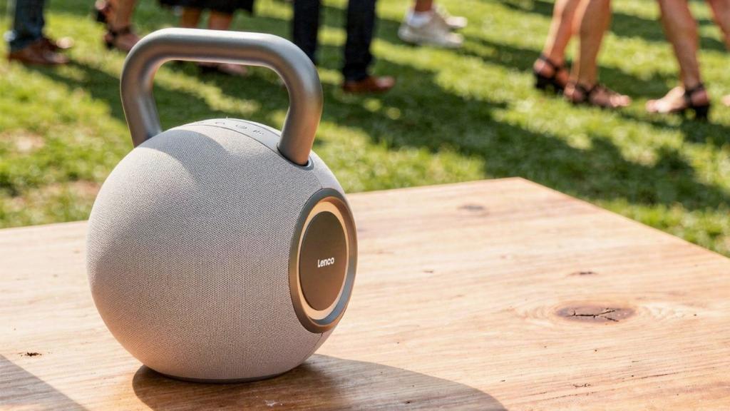 A kettlebell-shaped Lenco Orbita-360 Bluetooth speaker on a wooden table. There are people standing on grass in the background.