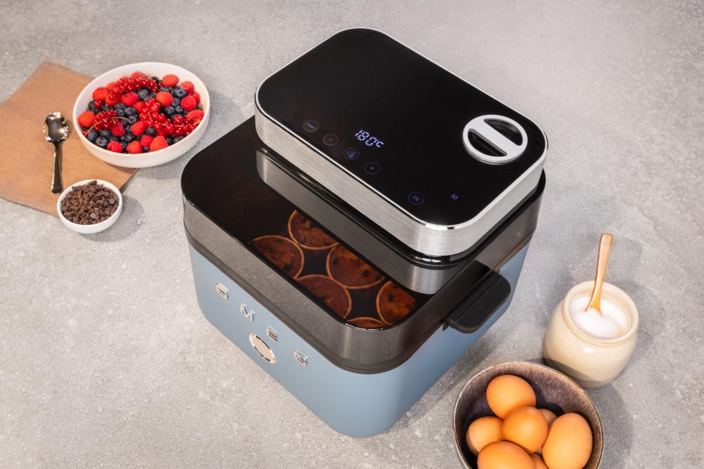 A blue Smeg air fryer on a grey surface. There is a bowl of fruit and a bowl of chocolate chips to the left of it, and a bowl of eggs and a bowl of sugar to the right.